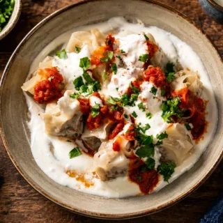 Steamed dumplings topped with creamy white sauce, red sauce, and chopped fresh herbs on a rustic table.